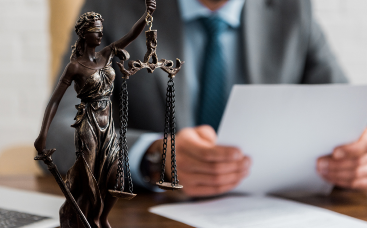What is the Difference Between a Defense Attorney and a Prosecutor? - scales of justice in the foreground with man in suit sitting at table holding paper in hands in the background